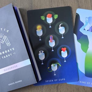 May include: A Mystic Mondays Tarot deck with a guidebook. The guidebook is gray with the text "Mystic Mondays Tarot Guidebook" and the author's name. Two tarot cards are visible, one labeled "Seven of Cups" and the other "King of Cups".