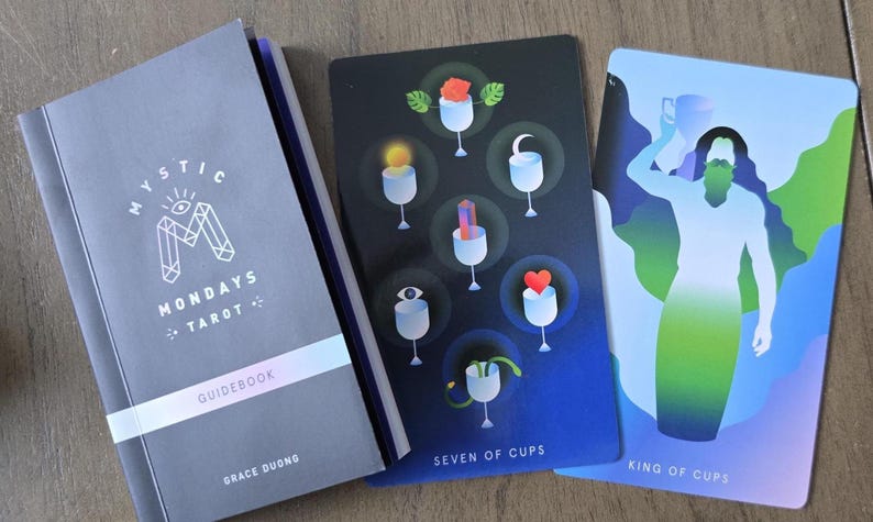 May include: A Mystic Mondays Tarot deck with a guidebook. The guidebook is gray with the text "Mystic Mondays Tarot Guidebook" and the author's name. Two tarot cards are visible, one labeled "Seven of Cups" and the other "King of Cups".
