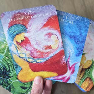May include: Five oracle cards with colorful illustrations and text. The cards are arranged in a fan shape. The text on the cards reads "Support", "Return to Innocence", "Breathe", "Presence", and "Presence".