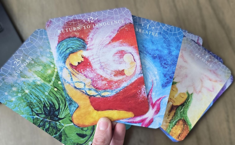 May include: Five oracle cards with colorful illustrations and text. The cards are arranged in a fan shape. The text on the cards reads "Support", "Return to Innocence", "Breathe", "Presence", and "Presence".