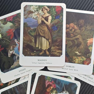 May include: A collection of tarot cards featuring illustrations of figures and scenes. The central card displays a woman labeled "Maiden." Other cards include "Goblin," "Steed," and "Sorcerer." The cards have white borders and are arranged on a dark surface.