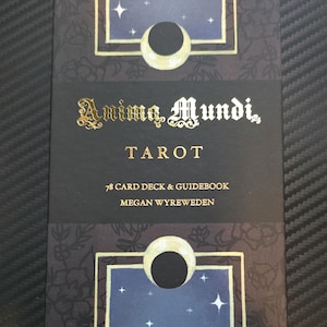 May include: A tarot card deck box with the title "Anima Mundi, Tarot". The box is black with gold lettering and features a celestial design with moons and stars. The box also includes the text "78 Card Deck & Guidebook" and the author's name.