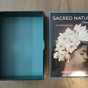 May include: A black box with a teal blue interior and a guidebook for the Sacred Nature Oracle deck. The guidebook cover features a young girl with a crown of white flowers on her head. The text on the cover reads "Sacred Nature Oracle" and "Guidebook".