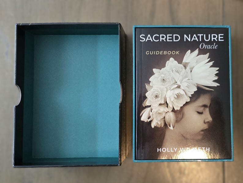 May include: A black box with a teal blue interior and a guidebook for the Sacred Nature Oracle deck. The guidebook cover features a young girl with a crown of white flowers on her head. The text on the cover reads "Sacred Nature Oracle" and "Guidebook".