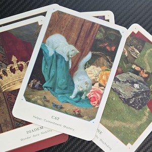 May include: A collection of tarot cards featuring illustrations of a golden crown, white cats, and floral arrangements. The cards have white borders and are stacked on a red book. The card with the cats is labeled "CAT".
