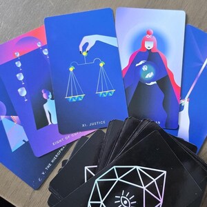 May include: A deck of tarot cards with vibrant illustrations. Several cards are fanned out, displaying images of scales, a figure with a globe, and other symbolic designs. The cards have a dark background with geometric patterns.