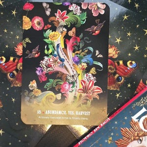 May include: A deck of tarot cards with a floral and butterfly design. The central card features colorful flowers, butterflies, and birds against a black background. The text "18 - ABUNDANCE, YES, HARVEST" is visible. Other cards have a starry background.