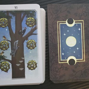 May include: A tarot card deck featuring the "VI" card, depicting a tree with golden pentacles, birds, and a squirrel. The card's reverse side has a blue background with stars, a moon, and floral accents.