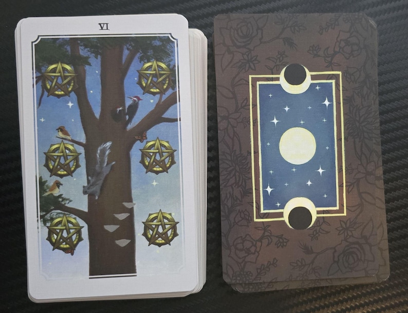 May include: A tarot card deck featuring the "VI" card, depicting a tree with golden pentacles, birds, and a squirrel. The card's reverse side has a blue background with stars, a moon, and floral accents.
