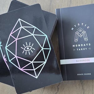 May include: A black tarot deck with a geometric design and an eye symbol, alongside a matching guidebook. The guidebook cover reads "Mystic Mondays Tarot" with the author's name. The cards have a geometric design with a pastel gradient.