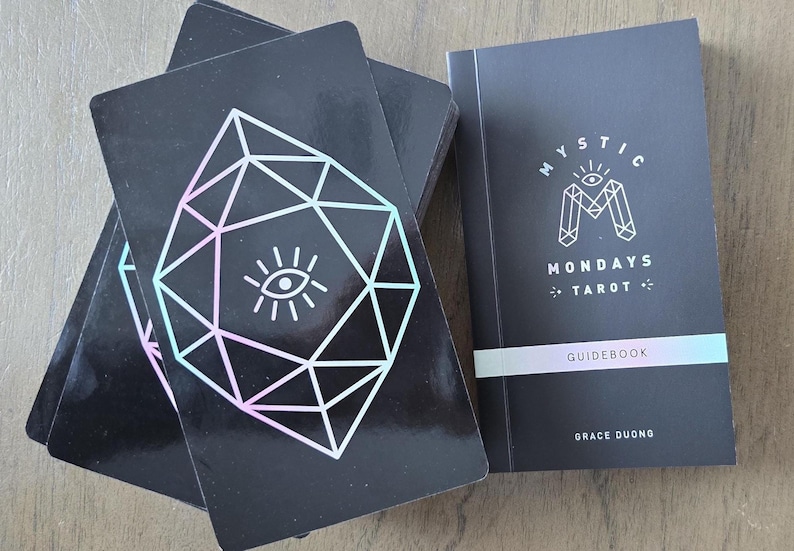 May include: A black tarot deck with a geometric design and an eye symbol, alongside a matching guidebook. The guidebook cover reads "Mystic Mondays Tarot" with the author's name. The cards have a geometric design with a pastel gradient.