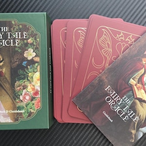 May include: A green box and a deck of red cards with a guidebook. The box reads "The Fairy Tale Oracle" with an image of a woman in a long dress. The cards and guidebook feature illustrations of fantasy scenes.