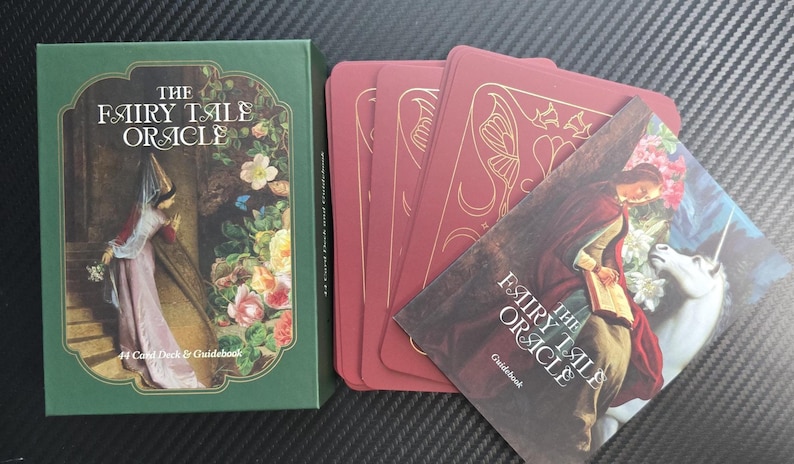 May include: A green box and a deck of red cards with a guidebook. The box reads "The Fairy Tale Oracle" with an image of a woman in a long dress. The cards and guidebook feature illustrations of fantasy scenes.
