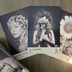 May include: Three oracle cards with black backgrounds and white text. The first card shows a woman with a floral crown and the text "HEART OPEN". The second card shows a woman with a floral headdress and the text "ALIGNMENT". The third card shows a sunflower and the text "FLOW".
