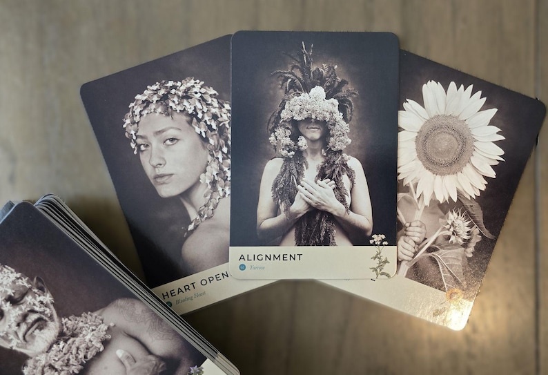 May include: Three oracle cards with black backgrounds and white text. The first card shows a woman with a floral crown and the text "HEART OPEN". The second card shows a woman with a floral headdress and the text "ALIGNMENT". The third card shows a sunflower and the text "FLOW".