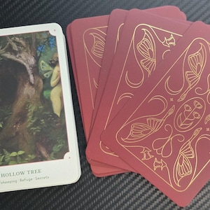 May include: A deck of tarot cards featuring a white card with a nature scene and the words "Hollow Tree." The other cards are burgundy with gold butterfly and floral designs.