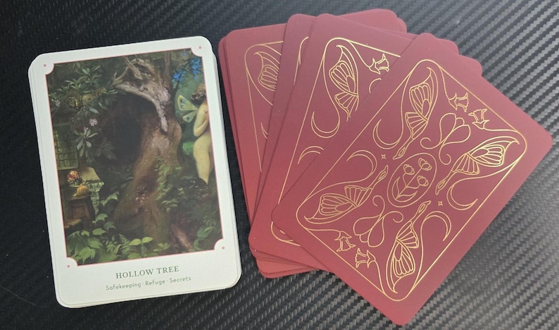 May include: A deck of tarot cards featuring a white card with a nature scene and the words "Hollow Tree." The other cards are burgundy with gold butterfly and floral designs.