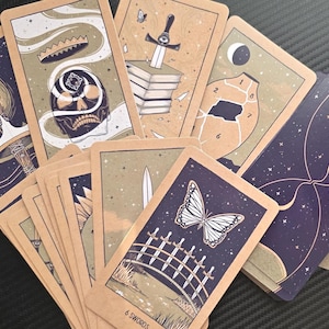 May include: A collection of tarot cards with unique illustrations. The cards feature images of a skull, a sword, a butterfly, and an hourglass, with a color palette of beige, navy blue, and white. The cards are arranged in a fan shape.