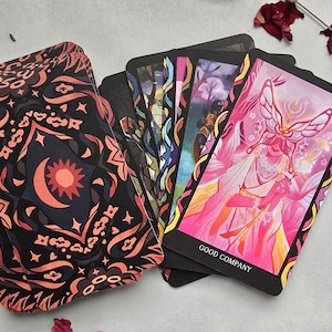 May include: A tarot card deck with a black and coral patterned box featuring a sun and moon design. The cards are fanned out, displaying colorful illustrations, including a card with a butterfly figure and the words "GOOD COMPANY".