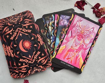 May include: A tarot card deck with a black and coral patterned box featuring a sun and moon design. The cards are fanned out, displaying colorful illustrations, including a card with a butterfly figure and the words "GOOD COMPANY".