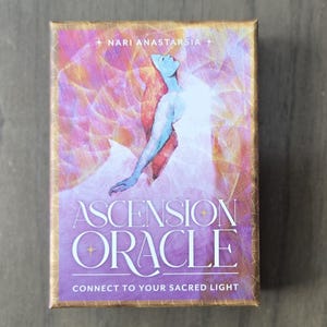 May include: A purple and gold box with the text "Ascension Oracle" and "Connect to Your Sacred Light". The box features a watercolor illustration of a woman with flowing red hair, wearing a white dress, with a pink and gold background.