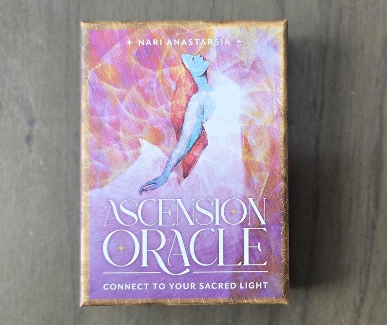 May include: A purple and gold box with the text "Ascension Oracle" and "Connect to Your Sacred Light". The box features a watercolor illustration of a woman with flowing red hair, wearing a white dress, with a pink and gold background.