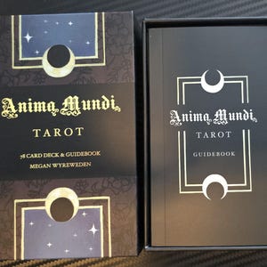 May include: A black tarot card box and guidebook set. The box features the text "Anima Mundi Tarot" in gold script, with a celestial design. The guidebook inside also displays the same title in white.