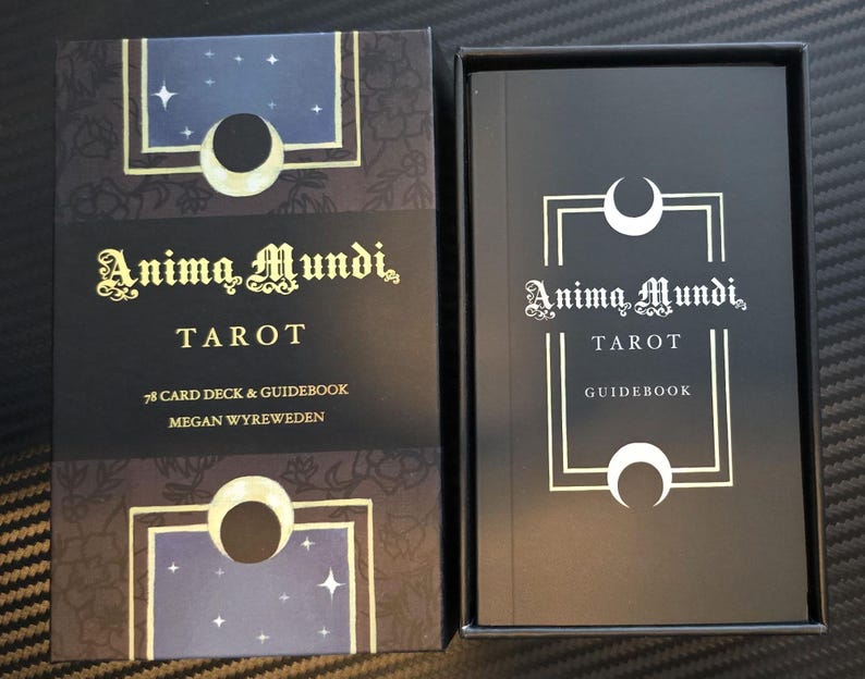 May include: A black tarot card box and guidebook set. The box features the text "Anima Mundi Tarot" in gold script, with a celestial design. The guidebook inside also displays the same title in white.