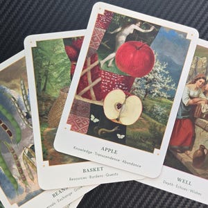 May include: A collection of tarot cards with various illustrations. One card features a red apple and the word "APPLE". Other cards depict images of beans, a basket, and two women. The cards have white borders and are arranged on a dark surface.