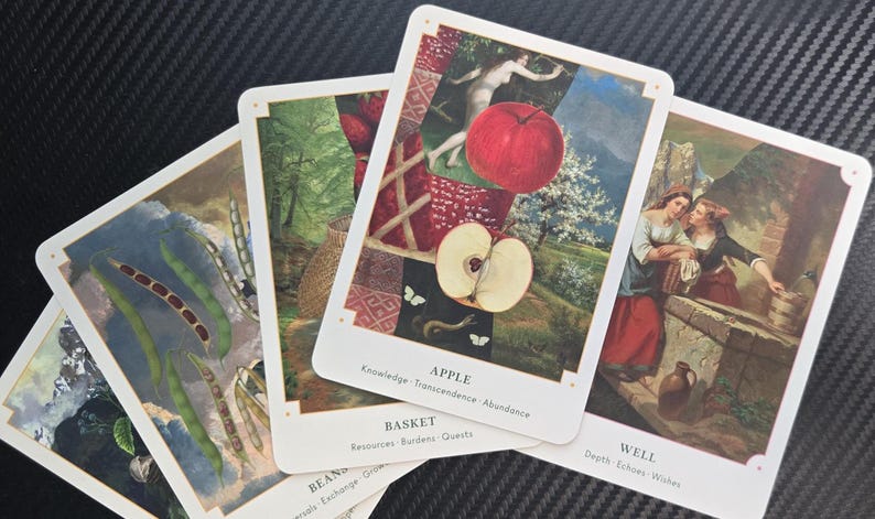 May include: A collection of tarot cards with various illustrations. One card features a red apple and the word "APPLE". Other cards depict images of beans, a basket, and two women. The cards have white borders and are arranged on a dark surface.