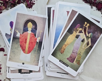 May include: A deck of tarot cards with various illustrations. One card features a figure in red robes and a gold hat, labeled "The Hierophant." Another shows three figures raising cups, labeled "Three of Cups."