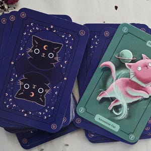 May include: A deck of tarot cards featuring a celestial cat theme. The card backs are navy blue with a cat illustration. One card shows a pink cat with a planet and the text "Ascension Cat" on a teal background.