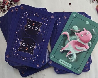 May include: A deck of tarot cards featuring a celestial cat theme. The card backs are navy blue with a cat illustration. One card shows a pink cat with a planet and the text "Ascension Cat" on a teal background.