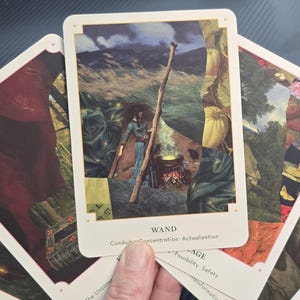 May include: A hand holding a deck of tarot cards. The central card features an illustration of a person with a staff near a fire, with the word "WAND" at the bottom. Other cards show various illustrations with white borders.