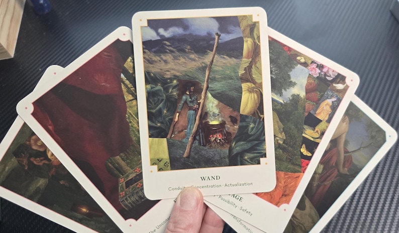 May include: A hand holding a deck of tarot cards. The central card features an illustration of a person with a staff near a fire, with the word "WAND" at the bottom. Other cards show various illustrations with white borders.