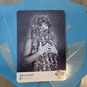 May include: A black and white tarot card with a person wearing a leafy headpiece and holding their hands to their chest. The card is titled "DELIGHT" with the text "#MagnificentYou" below the title.