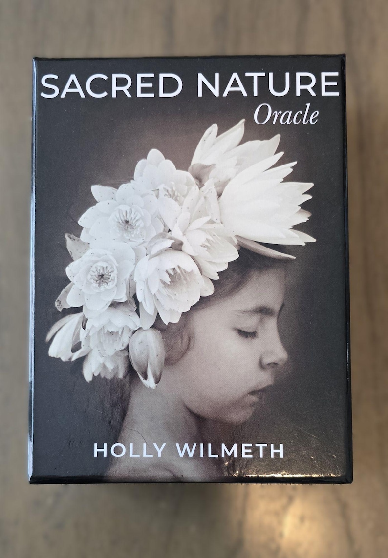 May include: A black box with the text "SACRED NATURE Oracle" and "HOLLY WILMETH" on the front. The box features a black and white photograph of a young girl with a crown of white flowers on her head.