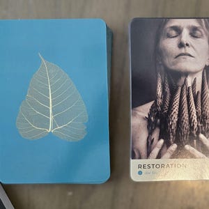 May include: A blue card with a white leaf skeleton on it. The card is part of a deck of cards called "Restoration" with the number 6 on the bottom left corner. The card on the right is a close-up of a woman with her eyes closed, holding a bouquet of flowers to her chest.