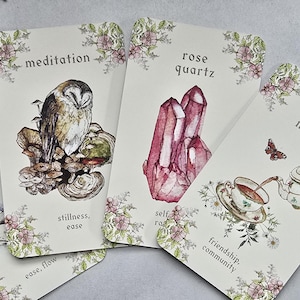 Puede incluir: Un conjunto de cartas de tarot con bordes florales e ilustraciones en acuarela. Las cartas presentan imágenes de un búho, cuarzo rosa, una tetera y una jarra de agua, con palabras como "meditación" y "magia del té".