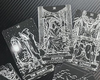 May include: Black and white tarot cards featuring detailed illustrations. The cards include "The Hermit," "Justice," and "The Lovers." The cards have a gothic aesthetic with intricate line art and celestial elements. The cards are displayed on a dark surface.