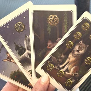 May include: A hand holding four tarot cards with illustrations of wolves, squirrels, and celestial elements. The cards feature gold pentacles and a night sky background. The cards are part of a tarot deck.