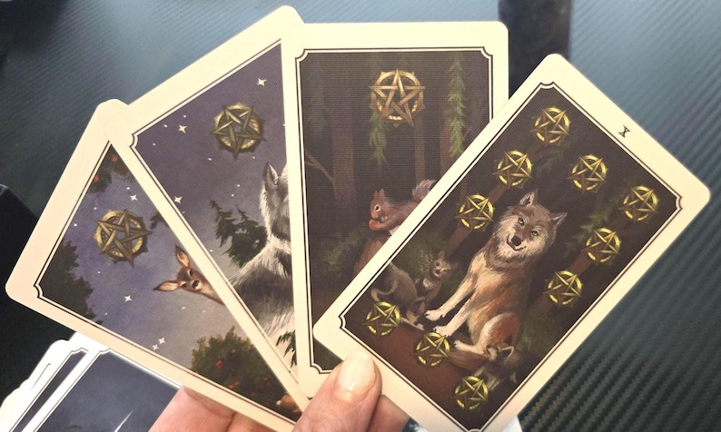 May include: A hand holding four tarot cards with illustrations of wolves, squirrels, and celestial elements. The cards feature gold pentacles and a night sky background. The cards are part of a tarot deck.