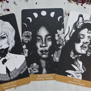 May include: Three tarot cards with black and white illustrations. One card features a woman holding a black cat, labeled "Curiosity." Another shows a woman with a crescent moon crown, labeled "Blessings." The third depicts a woman with butterflies, labeled "Wish."