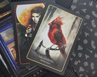 May include: A deck of oracle cards featuring illustrations. One card shows a red cardinal perched on a branch, labeled "NIGHTSONG" with "Hidden talents" below. Other cards depict a woman with a glass and a window. The card backs have a Halloween-themed pattern.