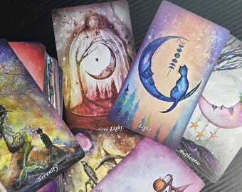 May include: A collection of tarot cards with celestial and mystical illustrations. The cards feature images of moons, cats, stars, and figures in a variety of colors, including blue, purple, and gold. Some cards have text such as "Light" and "Serenity".