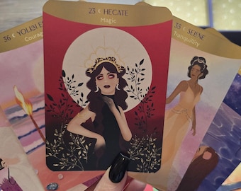 May include: A spread of tarot cards with gold accents and illustrations of people. The cards are laid out on a wooden surface. The cards include "8 C FUL MOON", "36 C YOUL BE", "25 C HECATE Magic", "35 C SERENE Tranquility", and "COMING MOON".