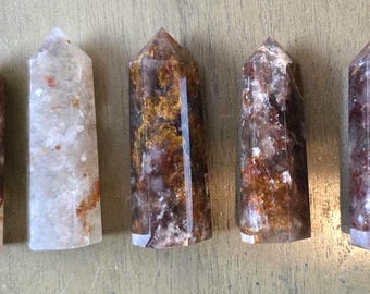 May include: Five crystal points with varying patterns and colors. The crystals are primarily shades of brown, red, and white, with a polished, smooth surface. Each crystal has a pointed top and a rectangular base. The crystals are arranged side by side.
