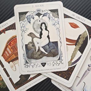 Könnte beinhalten: Eine Sammlung von Tarotkarten mit verschiedenen Illustrationen. Die zentrale Karte, mit der Aufschrift "2. Selkie", zeigt eine Frau, die auf einem Seehund sitzt. Weitere Karten zeigen Bilder eines Wohnwagens, einer Schere und einer stilisierten Lippe. Die Karten liegen auf einer dunklen Oberfläche.