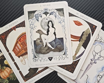 Op de afbeelding: Een verzameling tarotkaarten met diverse illustraties. De centrale kaart, met het label "2. Selkie", toont een vrouw zittend op een zeehond. Andere kaarten tonen afbeeldingen van een caravan, een schaar en een gestileerde lip. De kaarten liggen op een donkere ondergrond.