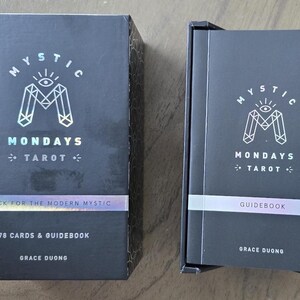 May include: Two black boxes of "Mystic Mondays Tarot" cards and guidebook. The boxes have a holographic logo and text that reads "A Deck for the Modern Mystic" and "78 Cards & Guidebook". The author's name, Grace Duong, is also printed on the boxes.